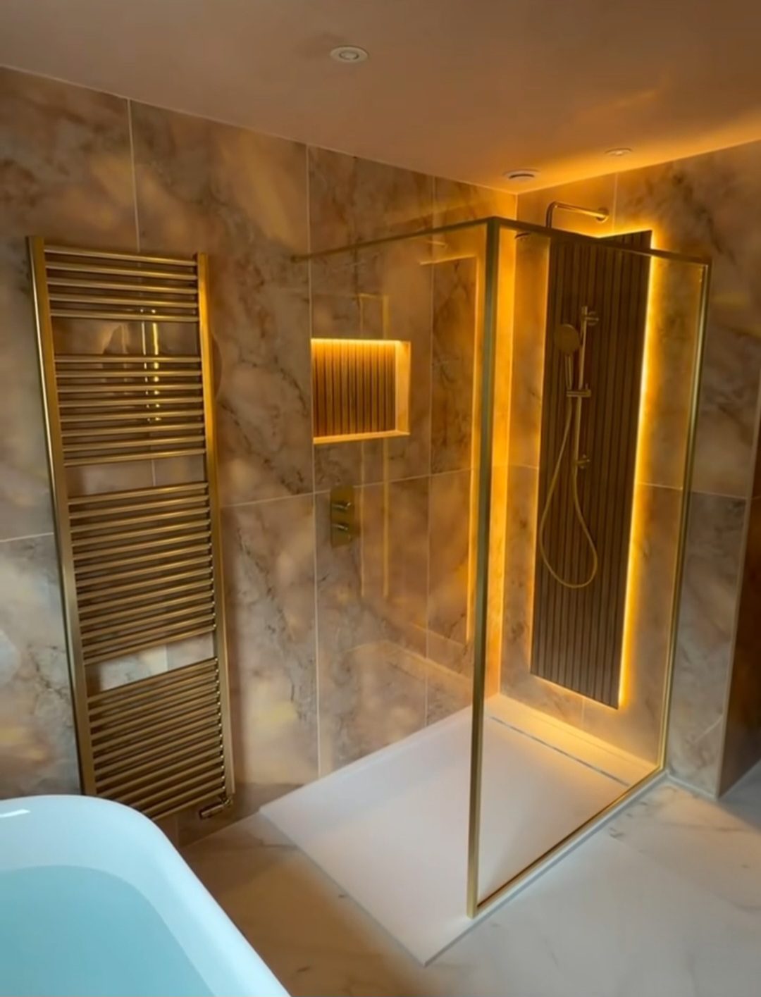 Walk-in shower with brushed gold frame, warm marble-effect tiling, and backlit niche