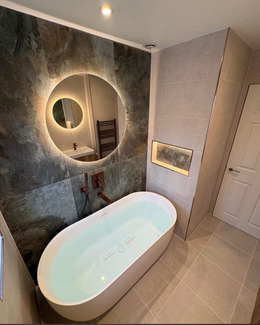 Freestanding bath beneath a backlit circular mirror, with natural stone-effect tiling and bronze fixtures